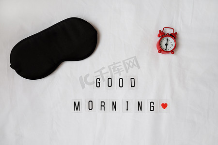 丝绸黑色睡眠面罩，题词早上好，白色皱巴巴的床单上的红色时钟。