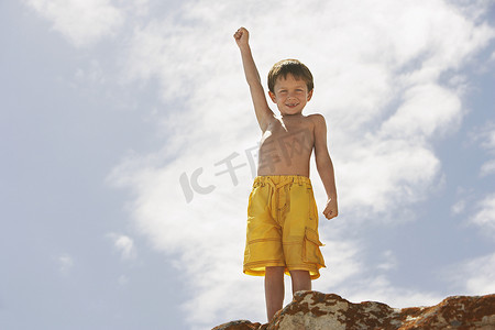 举手小男孩摄影照片_多云天空中举手站在岩石上的小男孩的低视角
