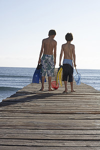 两个赤裸上身的男孩带着渔网和泳蹼在码头上行走的全长后视图