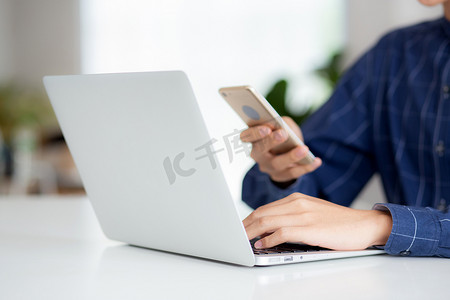 年轻人在家里的办公桌上在线工作笔记本电脑和阅读智能手机的特写手，自由职业男性使用手机与社交媒体、商业和通信概念。