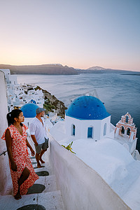 希腊圣托里尼岛，年轻夫妇在圣托里尼岛豪华度假，在日出期间在蓝色圆顶教堂和希腊伊亚圣托里尼粉刷成白色的村庄旁观看日出，男女在希腊度假