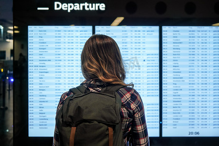 身穿绿色背包和衬衫的年轻女子看着机场的出发板屏幕，从后面看