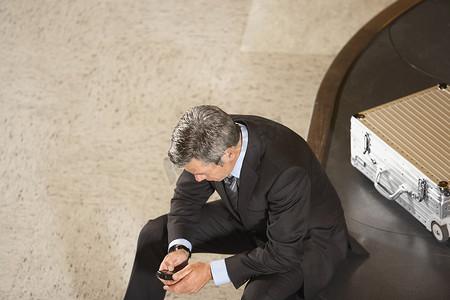 一位商人坐在机场转盘上的行李旁看手机的高视角