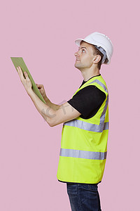 一名男性建筑工人的侧视图，带有剪贴板，在粉红色背景下望向别处