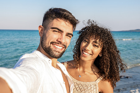 黄昏时分，美丽的混血夫妇在海边自拍 — 穿着白色开衫的英俊年轻人和他的拉丁卷发女友在暑假里一起微笑