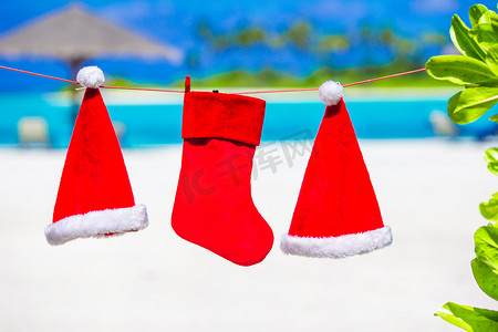 挂圣诞袜摄影照片_挂在热带海滩上的红色圣诞老人帽子和圣诞袜