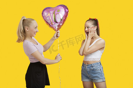 快乐的年轻女人给站在黄色背景下惊讶的女人送生日气球
