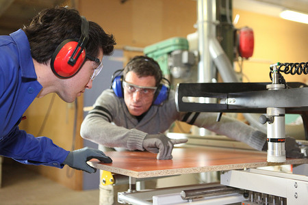 学徒制摄影照片_一位经验丰富的工人向学徒展示如何使用机器切割一块木头