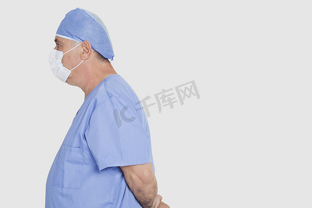 灰色背景下双手背后站立的高级男性外科医生的侧面照片
