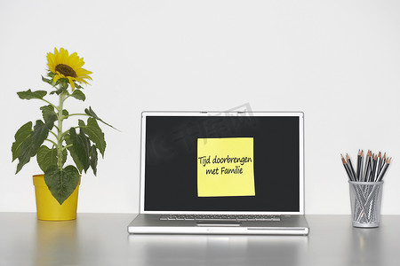 桌上的向日葵植物和笔记本电脑屏幕上贴着荷兰语文字的便签纸，上面写着“Tijd Doorbrengen met Familie”（与家人共度时光）