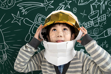 儿童宇宙摄影照片_戴着航天员头盔的小男孩站在黑板前