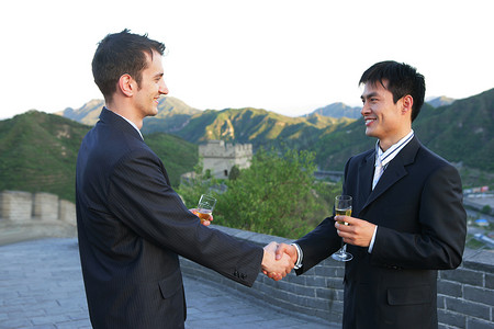 两位中外商务人士在长城上端酒杯握手