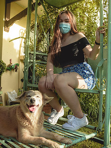一个戴着面具的快乐的年轻亚洲女人和她忠实的狗坐在滑翔机的秋千上。新的正常概念.