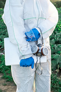 一位穿着白色防护服、面罩和蓝色手套的农艺学家，带着收获把笔记本电脑顶在田边。化学品和农药概念问题.
