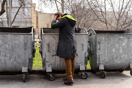 垃圾箱和摄影照片_绝望的年轻人抬起头，在垃圾箱里寻找食物。一个在垃圾箱里找食物而失业的孤独男人。无家可归和失业的人的问题。街头流浪者的生活方式