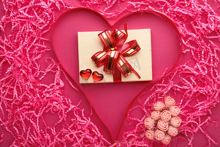 粉色彩带背景摄影照片_在粉色的背景上，有一个巨大的心，由纸张、卵石、彩带和一束玫瑰做成，装饰在礼物里面.