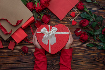 海报2摄影照片_女性的手捧着心形的盒子，情人的礼物在木制的表面，玫瑰，花瓣，红色的购物袋。情人节2月14日或浪漫的夜晚准备工作、明信片、海报、装饰品