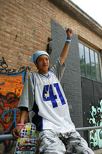 拿滑板的男青年坐在798艺术区内的涂鸦画前