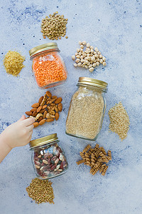 Cereal grains, seeds, beans in mason jars on a blue background. Child's hand touch takes nuts, healthy food concept