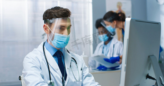 年轻英俊的白种人男医生头戴口罩坐在橱柜里的近照，在医疗中心用计算机进行检疫打字和检查医生的概念时进行面罩工作