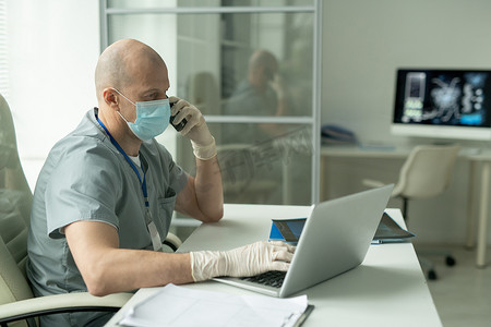狗熊侧影摄影照片_在医务室的笔记本电脑前，坐在桌子旁，一边用手机交谈，一边看戴防护面具的秃头男性临床医生的侧影