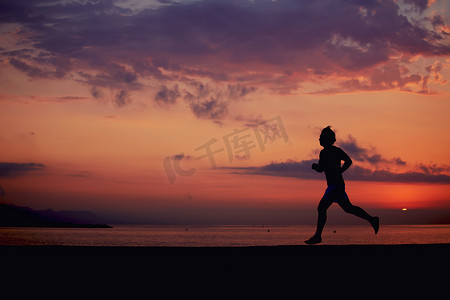海滩晚霞摄影照片_肌肉健美运动员的轮廓在海滩上跑得很快，跑步者在海面上迎着五彩缤纷的日出慢跑，男性慢跑者身强体壮地奔跑