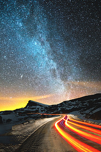 夜风景。夜空中有一颗北半球的银河和星星。由汽车风照亮的夜路与一条蛇形和叶子在距离到一英尺的尖锐岩石。光径