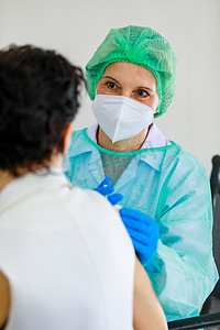 白种人老年女医生身穿PPE全副危险防护服，头戴口罩，在医院接种台为妇女肩部注射疫苗.
