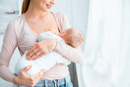 裁剪拍摄微笑的年轻母亲站在和母乳喂养婴儿在家里