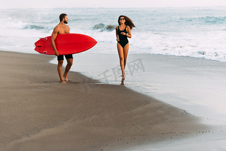 在海滩上和冲浪板在夕阳下坠入爱河。巴厘岛的一对年轻漂亮的夫妇。健康生活方式的概念。一对夫妇冲浪者走在印度尼西亚的海岸上。在海上度假