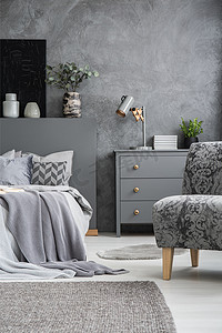单色灰色卧室内有一张床、一个抽屉柜、一盏台灯、一把扶手椅、植物和花瓶。地板上的地毯。在质感的墙壁上的黑色绘画。真实照片.
