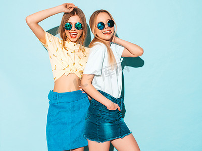 两个年轻美丽的微笑的金发嬉皮士女孩在时尚的夏季牛仔裤裙子衣服。性感无忧无虑的妇女摆出近蓝色墙壁在圆形太阳镜。积极的模型, 有乐趣, 并显示和平标志