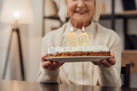 裁剪视图微笑的高级妇女拿着生日蛋糕与数字80在家里 
