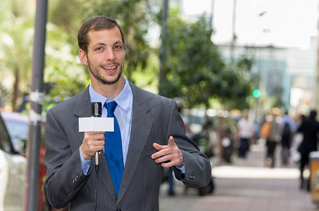 Attractive professional male news reporter wearing grey suit holding microphone, talking to camera from urban setting