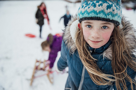 小女孩正在看着相机, 同时拉着雪橇上山在雪中与她的朋友.