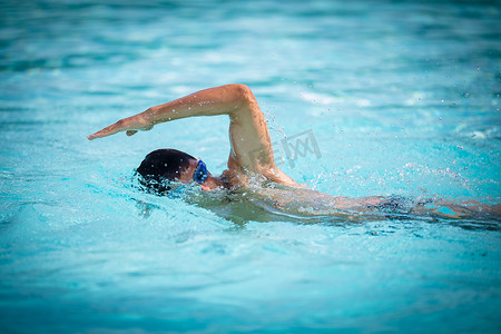 游泳泳镜摄影照片_男子游泳者在蓝色的游泳池里爬泳.一名身穿泳镜的年轻男子游泳爬行运动员的肖像。铁人三项全能运动员训练.