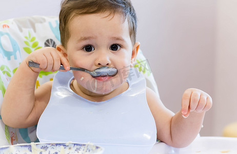 微笑圆形表情包摄影照片_微笑的孩子在厨房吃的食物。1 年岁宝宝吃他们自己的汤。第一次的固体食物，如何教孩子用勺子吃饭。可爱的宝宝，吃午饭用勺子吃饭