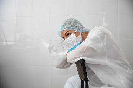 口罩帽子摄影照片_一位身穿防护服、头戴帽子、戴着面具、戴着手套的年轻女医生疲惫不堪地坐在医务室的椅子上