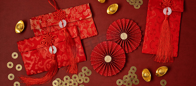 Chinese new year festival decoration over red background. Traditional lunar new year red pockets, paper fans, coins, gold ingots with text meaning fortune, prosperity, wealth. Flat lay, top view, banner