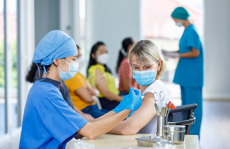 亚洲女医生头戴面罩，身穿蓝色医院制服，为白种人老年妇女注射疫苗，并配备设备，而其他病人则在模糊的背景下排队等候.