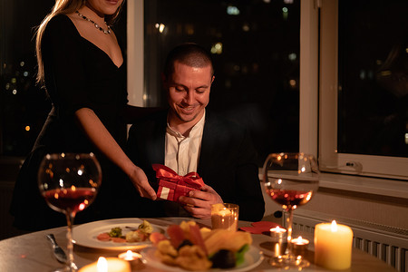 情人节那天晚上，一个女孩用蜡烛送礼物给一个男人晚餐，两人共进晚餐，一个装有惊喜礼物的盒子。情人节庆祝的概念。横向射击.