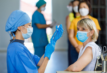 可爱的医生戴着面罩，戴着橡胶手套，身穿蓝色医院制服，在白人女性病人等待接种疫苗时，用针头注射器从小玻璃瓶中吸出考拉威斯疫苗.