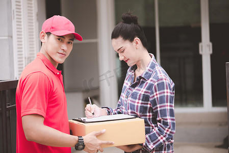 年轻的后勤职业概念。快乐送货的人把他的包给客户在家里。在真正的房子。二十岁代初, 亚洲华人身穿红色马球衫和红帽牛仔裤.