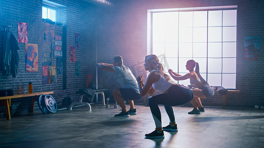 强壮的男子男子和两名健康的女子正在做蹲姿练习。他们在带激励海报的阁楼健身房里锻炼。有洋红色灯的房间。他们团队精神的一部分