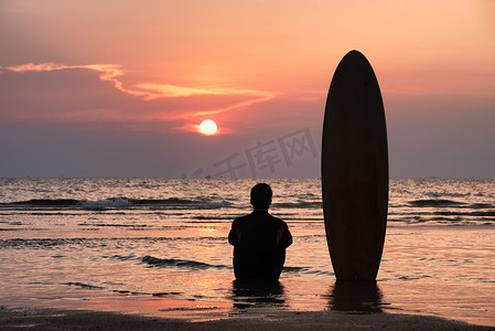 沙滩冲浪板摄影照片_冲浪者独自坐在沙滩上冲浪后，在日落时分用冲浪板眺望大海