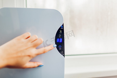 女子在湿窗使用触摸屏时转动除湿机。湿度指示器显示平面的湿度较高。空气干燥机