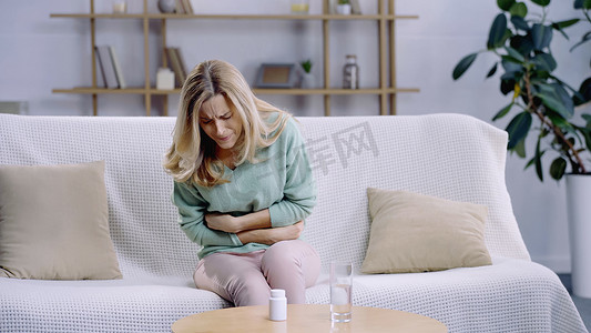 坐在茶几上的一杯水和药丸旁边的金发女人肚子痛 