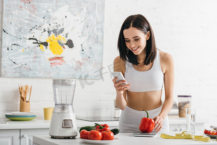 Smiling sportswoman using smartphone near scales, vegetables and measuring tape on kitchen table, calorie counting diet
