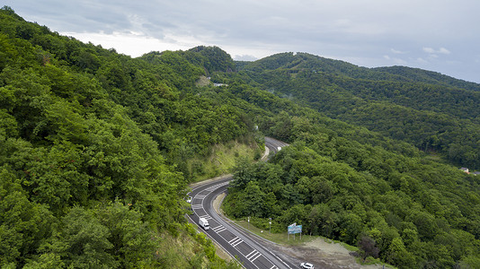 祈福之旅摄影照片_在俄罗斯索契的森林里, 沿着蜿蜒的山路行驶的汽车的空中股票照片。人行, 路之旅, 穿越美丽的乡村风光.