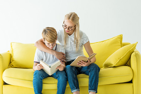 母亲在眼镜拥抱儿子, 而他读的书上的黄色沙发上孤立的白色